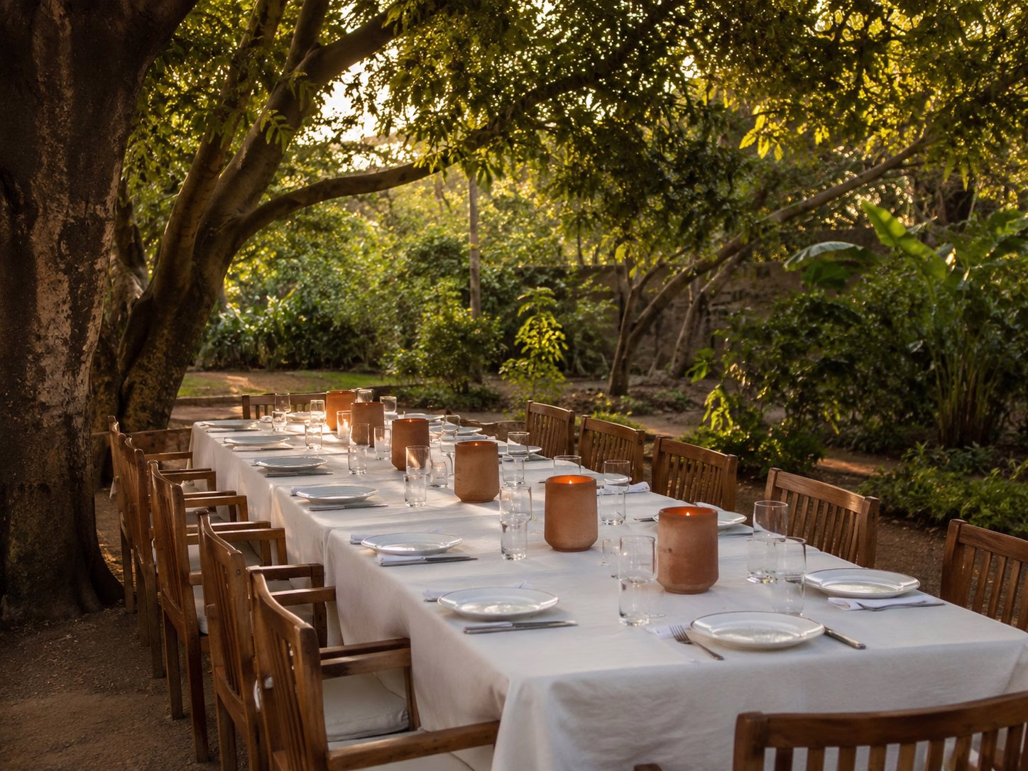 Long outdoor dining table set under tropical trees with clay candle holders, golden light
