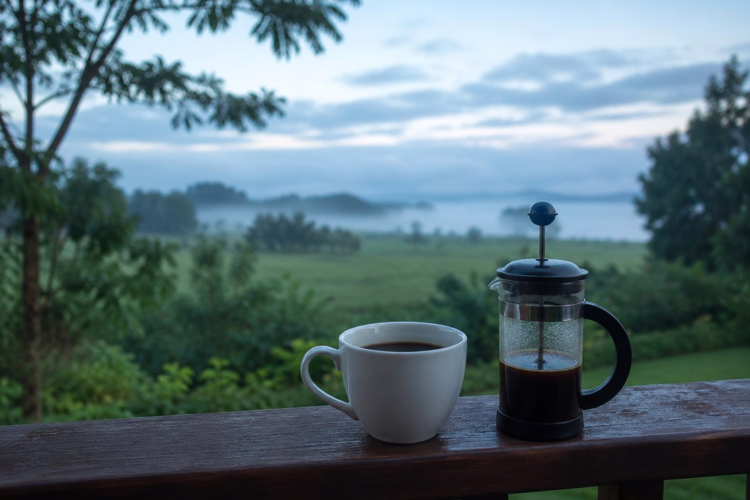 White ceramic mug and french press on a wooden veranda railing at dawn with misty lake view